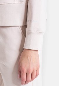 Jasnoróżowy sweter z ribbowanymi mankietami i miękką fakturą. Posiada haftowany napis na rękawie. Zbliżenie na ramię i dłoń.