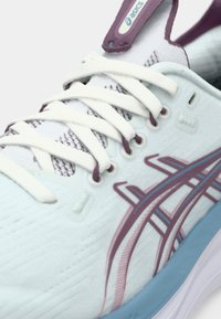 Světle modrá sportovní obuv s bílými tkaničkami, fialovými akcenty a texturovanou tkaninou. Má zakřivený tvar a výrazné logo značky.