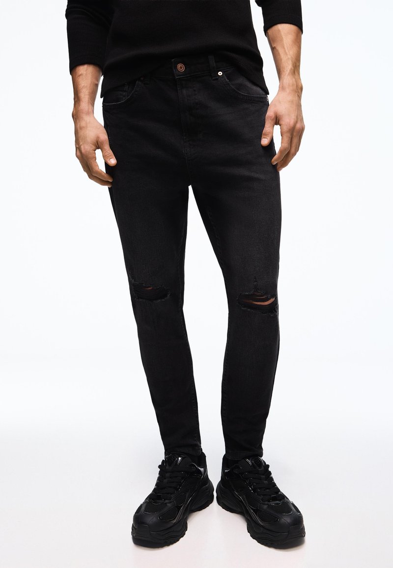 Férfi vékony fekete, szakadt farmernadrágban, fekete masszív tornacipőben és fekete hosszú ujjú felsőben, fehér háttér előtt állva.