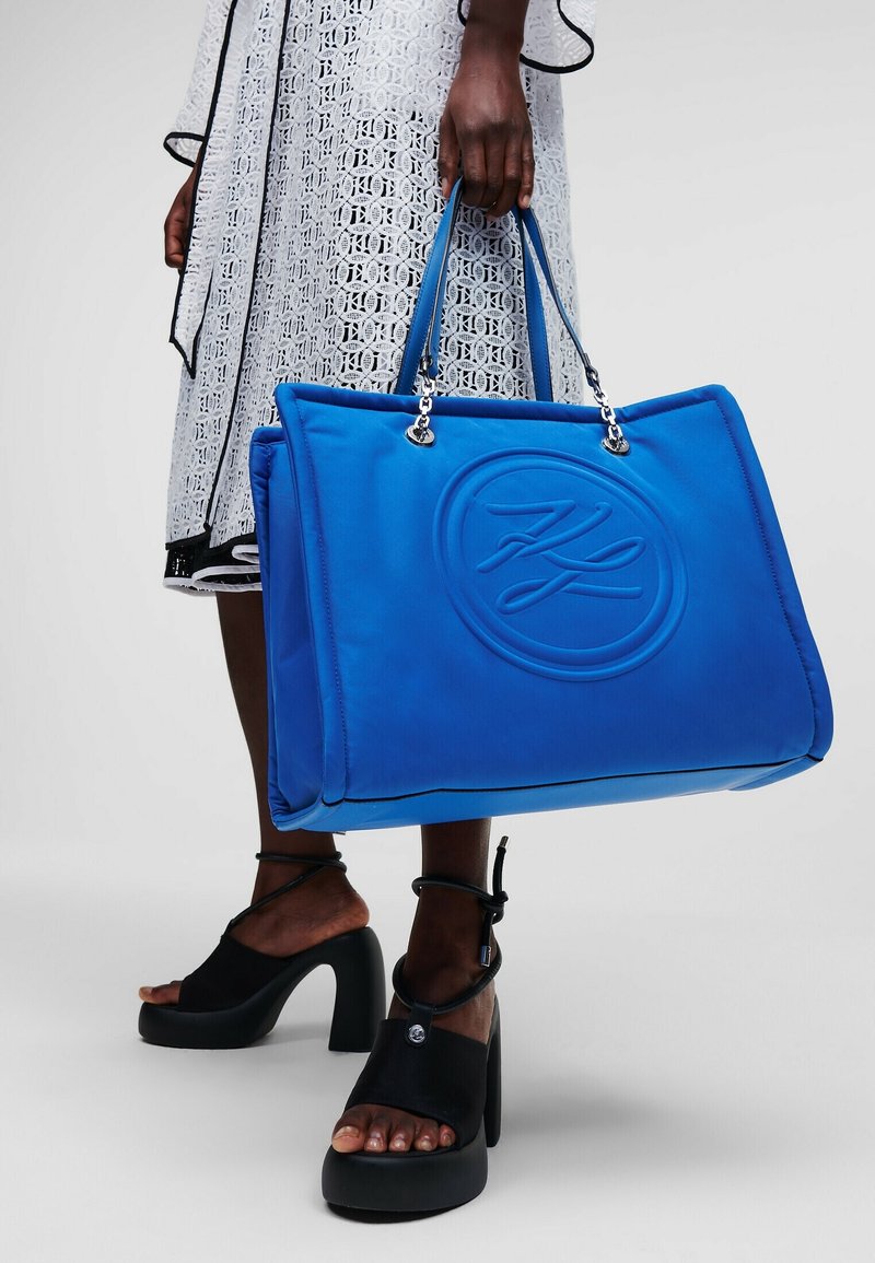 Personne tenant un grand sac à main bleu vif avec logo en relief, portant des sandales à plateforme noires et une robe blanche à motifs.