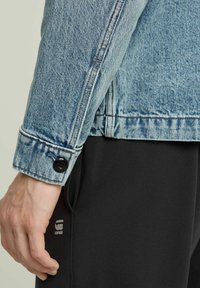 Ljusblå denimjacka med knappärmar, kontraststickningar och en avslappnad passform, i kombination med svarta byxor som visar en logotyp.