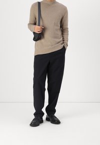 Person trägt einen beigen strukturierten Pullover, schwarze lässige Hosen, schwarze Schuhe und hält eine schwarze Umhängetasche über der Schulter.