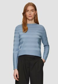 Frau mit roten Haaren, die einen hellblauen gestreiften Langarmpullover und schwarze Hosen trägt, steht mit einer Hand in der Tasche vor einem schlichten Hintergrund.