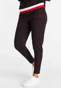 Pantalones de rayas negras y rojas con una silueta ajustada, combinados con zapatillas blancas, que presentan un logo decorativo en la cadera y una cinturilla en contraste.