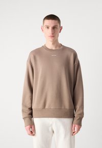 Hnedý crewneck sveter s rebrovanými manžetami a lemom, s malým bielym logom "Calvin Klein" na hrudi. Hladká textúra látky.