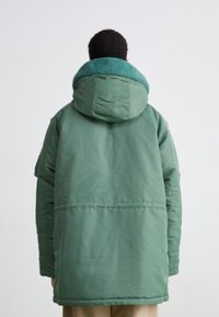 Grüne Steppjacke mit einem türkisfarbenen, flauschigen Kragen. Verfügt über eine Kapuze und einen geraden Schnitt. Glatte Außenseite mit sichtbaren Steppnähten.