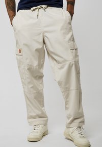 Pantalons cargo beiges en tissu léger, dotés d'une taille élastique à cordon de serrage, de poches latérales et d'un motif texturé discret. Associes à des baskets blanches.
