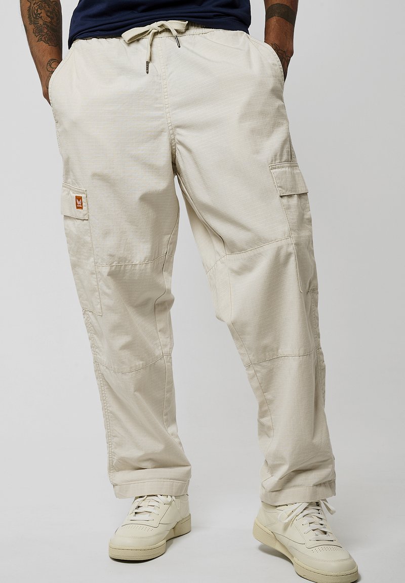 Pantalons cargo beiges en tissu léger, dotés d'une taille élastique à cordon de serrage, de poches latérales et d'un motif texturé discret. Associes à des baskets blanches.