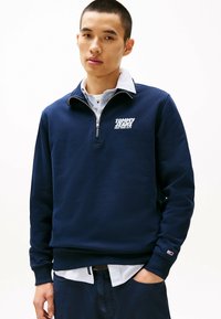 Navyblauwe zip-up hoodie met een wit logo op de borst, van katoen, met ribbels aan de mouwen, in laagjes over een lichtblauw overhemd.