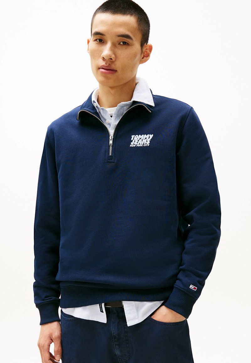 Navyblauwe zip-up hoodie met een wit logo op de borst, van katoen, met ribbels aan de mouwen, in laagjes over een lichtblauw overhemd.