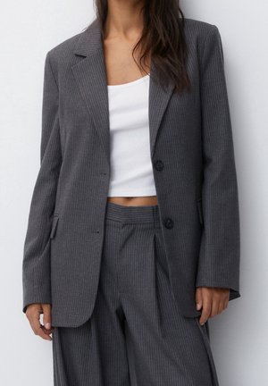Femme portant un costume gris foncé à fines rayures avec un top court blanc, debout devant un fond clair uni.