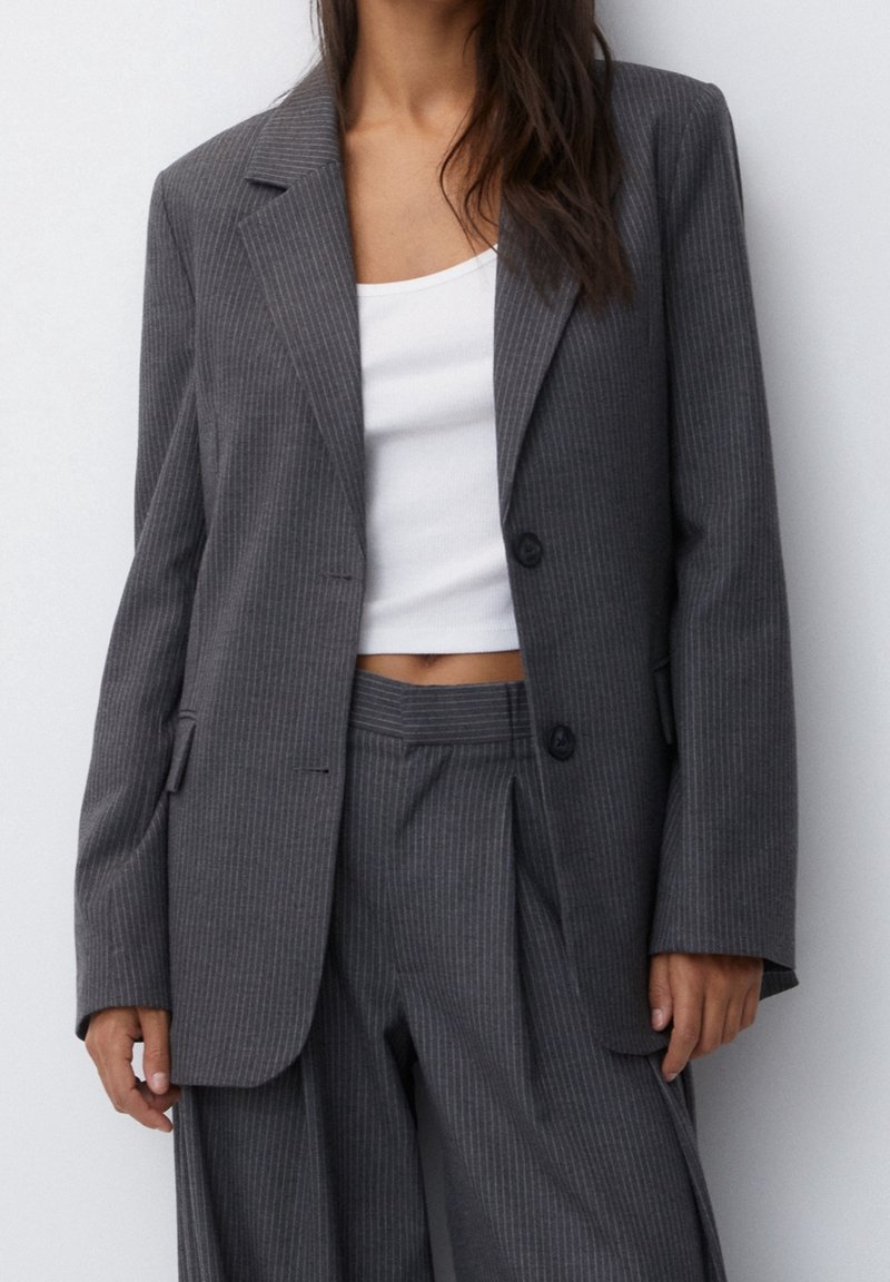 Femme portant un costume gris foncé à fines rayures avec un top court blanc, debout devant un fond clair uni.