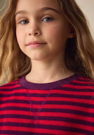 Mladé dievča s dlhými blond vlasmi, ktoré nosí červené a fialové pruhované tričko, pokojne hľadí dopredu na neutrálnom pozadí.