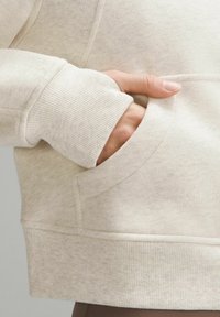 Ruka v přední kapse světle béžové mikiny s vroubkovaným manžetovým okrajem, zobrazení textury látky a detailů šití.