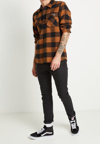 Ruskea ja musta ruudullinen flanellipaitaja, jossa on kaksi rintataskua, mustat housut ja mustat korkeat tennarit valkoisilla yksityiskohdilla ja nauhoilla.