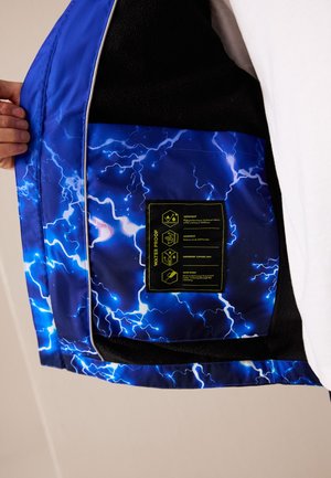 Blauwe jas binnenkant met bliksemschichtpatroon en zwart label dat waterdicht, winddicht en gesealde naden beschrijft, opengehouden door een hand.