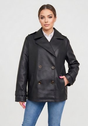 Młoda kobieta o neutralnym wyrazie twarzy, ubrana w czarną dwurzędową skórzaną kurtkę, białą koszulę i niebieskie dżinsy, stojąca na jednolitym tle.