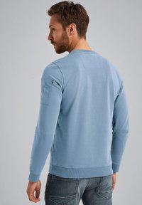 Sweatshirt bleu clair avec des manches longues, des poignets côtelés et un patch dans le dos ; texture douce, design décontracté et détail de poche latérale.