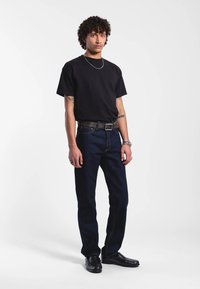 Musta pyöreäkauluksinen t-paita, tummansiniset suorat farkut, musta vyö metallisoljella ja mustat kengät; mallilla on lyhyet kiharat hiukset.