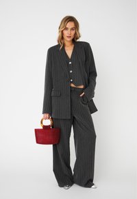 Costume gris à rayures avec blazer surdimensionné et pantalon large ; le mannequin tient un sac à main texturé rouge avec des poignées circulaires. Chaussures argentées visibles.