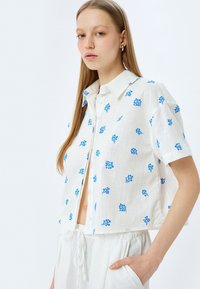 Camicetta bianca a maniche corte con stampa floreale blu, apertura con bottoni e design cropped, realizzata in tessuto leggero.