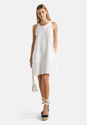 Vestido de lino blanco con un corte holgado, presenta un cuello redondo y bolsillos laterales. La modelo lleva sandalias de esparto negras con cuña y sostiene un bolso blanco.