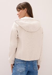 Veste en polaire gris clair avec capuche, présentant des poignets côtelés et une coupe courte. Texture douce, détails minimalistes et coupe décontractée.