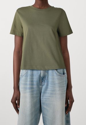 Oliwkowa koszulka z krótkim rękawem i okrągłym dekoltem, wykonana z gładkiej tkaniny, w zestawieniu z jasnoniebieskimi szerokimi jeansami z denimu.