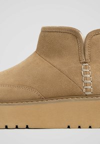 Bottine en daim marron avec une semelle plate. Présente des détails de couture et une doublure intérieure lisse pour le confort.