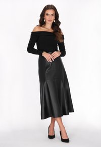 Donna che indossa un top nero a spalle scoperte, una gonna midi in pelle nera, tacchi alti neri e orecchini dorati, in piedi con le mani unite.