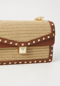 Beige geweven strohandtas met bruine leren accenten, gouden piramide-studs en een gouden sluiting. Gestructureerd ontwerp met een textuur oppervlakte.
