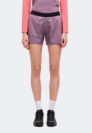 Persona che indossa pantaloncini viola e maglietta corallo a maniche lunghe, in piedi con le braccia rilassate, calzini bianchi e scarpe da ginnastica nere su sfondo chiaro.