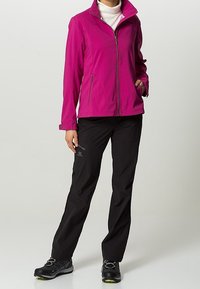 Persona vestida con una chaqueta de cremallera magenta, un suéter de cuello alto blanco, pantalones negros y zapatos de senderismo negros, de pie con una mano en el bolsillo.