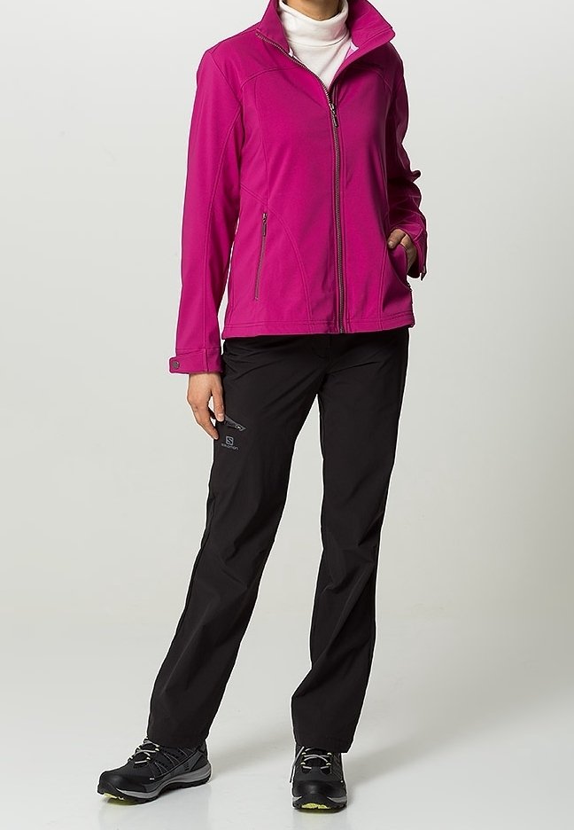 Persona vestida con una chaqueta de cremallera magenta, un suéter de cuello alto blanco, pantalones negros y zapatos de senderismo negros, de pie con una mano en el bolsillo.