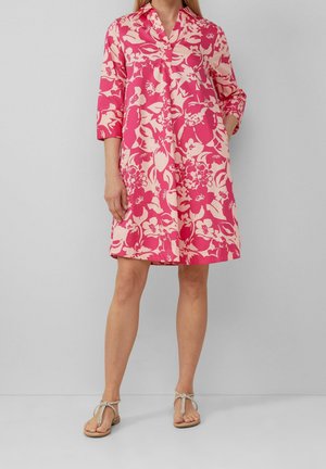 Kobieta ubrana w różowo-białą sukienkę o długości do kolan z kwiatowym wzorem, z rękawami do łokci, oraz beżowe płaskie sandały, stojąca na tle jasnoszarej ściany.