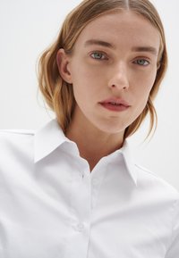 Lähikuva henkilöstä, jolla on vaalea iho ja vaaleanruskeat hiukset, valkoista kauluspaitaa käyttävä, katsoo suoraan eteenpäin neutraali ilme kasvoillaan.