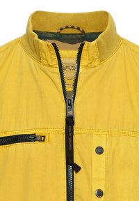 Gelbe Jacke mit Reißverschluss und Stehkragen, aus strukturiertem Stoff, mit kontrastierenden schwarzen Reißverschlüssen und einer Brusttasche.