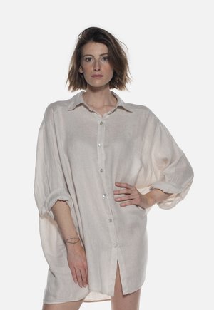 Donna con capelli corti e castani che indossa un vestito camicia oversize beige chiaro con bottoni, una mano sui fianchi e un’espressione neutra.