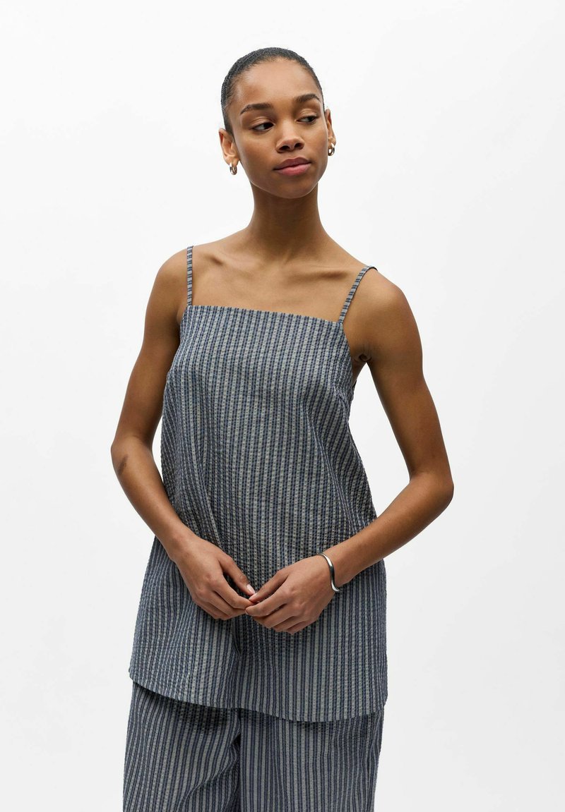 Jeune femme aux cheveux courts portant un haut sans manches rayé bleu-gris et un pantalon large assorti, debout sur un fond blanc.