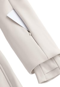 Jasnobeżowy rękaw kurtki z kieszenią na zamek, w której znajduje się biała karta. Miękka tekstura i smukły design.