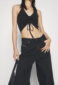 Black halter top with a ruching detail at the center and ties. Paired with high-waisted black denim wide-leg jeans featuring a colored logo accent.