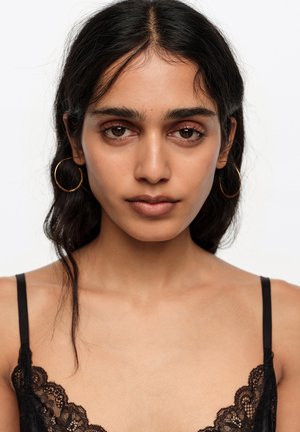 Joven mujer con cabello oscuro, lleva pendientes de aro grandes y una blusa negra con encaje, mirando directamente a la cámara sobre un fondo blanco.