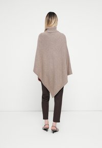 Poncho tricotado castanho claro com gola alta, apresentando uma silhueta triangular e uma superfície texturizada. Usado sobre calças castanhas escuras.