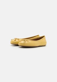 Un paio di ballerine gialle in pelle con punte rotonde e piccoli fiocchi decorativi su sfondo bianco.