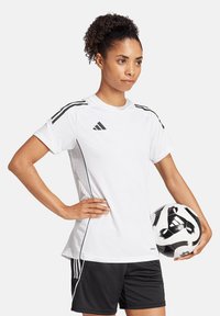 Maglietta sportiva bianca con bordini neri, maniche corte e tre strisce sulle spalle; pantaloni corti neri; tiene in mano un pallone da calcio bianco e nero.