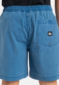 Shorts albaștri confecționați dintr-un material cu o textură netedă. Dispun de un brâu elastic și o buzunar la spate cu o etichetă de marcă.