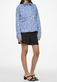 Farkkutakki vaaleansinisellä värillä, jossa on valkoista päivänkakkara-ompelua, metalliset napit, kaksi etutaskua ja rento malli. Yhdistetty mustiin shortseihin.