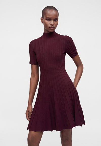 Femme portant une robe bordeaux côtelée à manches courtes, avec un col montant et une jupe plissée, debout devant un fond clair uni.