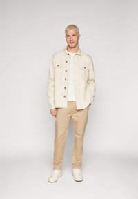 Veste beige à boutons sur une chemise blanche, chinos khaki et baskets blanches. Présente deux poches poitrine et un design décontracté.