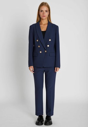 Kvinde iført navyblå dobbeltradet blazer med guldknapper og matchende bukser, står foran en ensfarvet lysegrå baggrund og vender fremad.